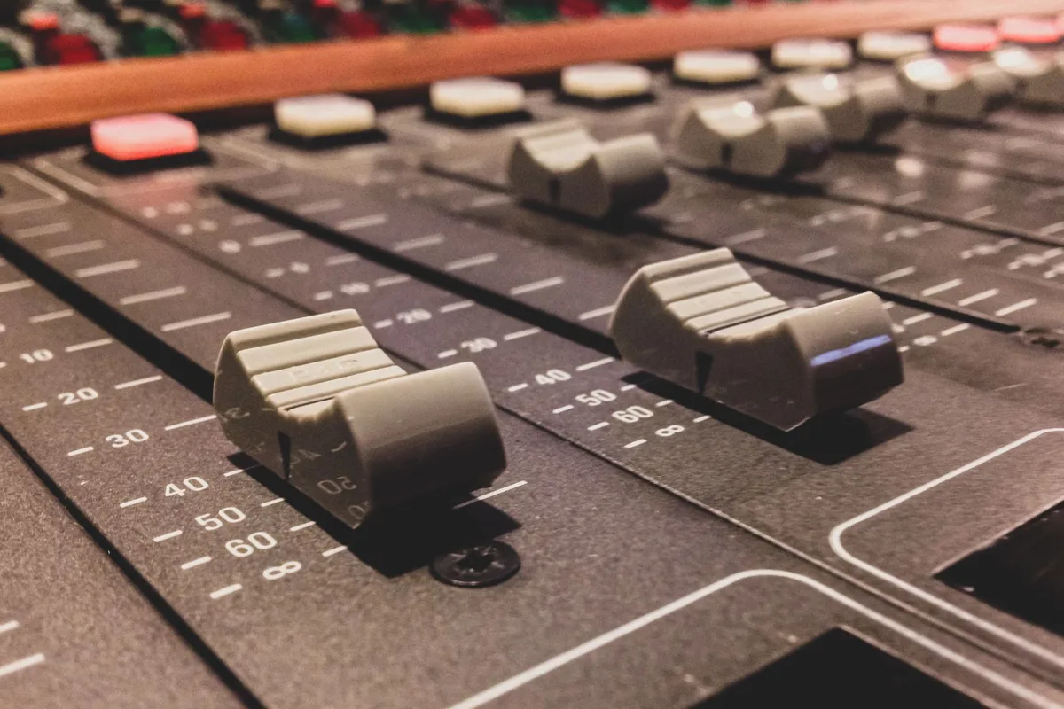 A mixing desk with faders in a darkened studio, lit from one side.